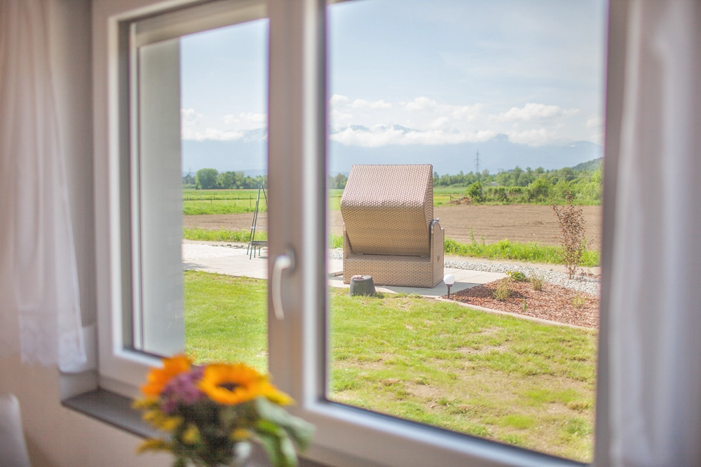 „Hahns Herberge“ – Raumaufnahme; gepflegte, helle Einrichtung, Blick durch das Fenster in den Garten, man sieht einen großen Liegestuhl und ein Feld im Hintergrund.